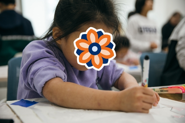 A young child sits at a desk drawing or writing, with her face covered by a flower sticker. Other children and adults are blurred in the background.
