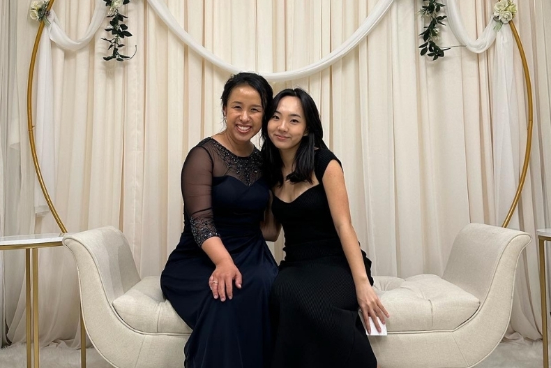 Two women dressed formally sit together on a cream-colored couch in front of an elegant backdrop with draped fabric and floral decorations, smiling at the camera.