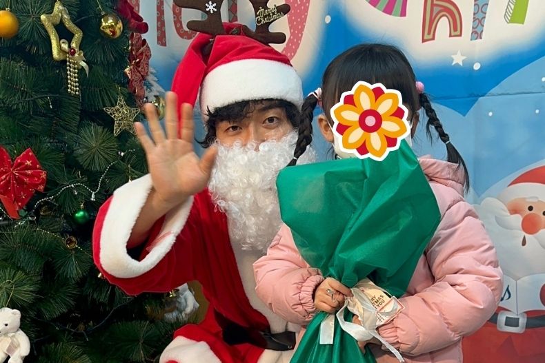 A volunteer dressed as Santa poses beside a young child holding a wrapped Christmas gift. The child’s face is covered with a flower sticker, and the backdrop features holiday decorations.