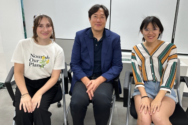 Two women sit on each side of a man in a blue blazer, all smiling at the camera. The women wear casual summer clothing, and the room has large windows in the background.