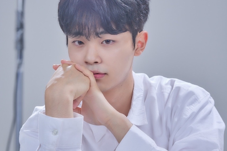 A young man in a white shirt poses for a portrait, resting his chin on his hands. He looks directly at the camera with a calm, thoughtful expression against a soft studio background.