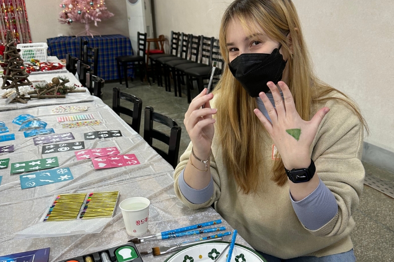 A volunteer wearing a black mask sits at a craft table holding a paintbrush and showing a small green painted shape on her hand. The table is covered with art supplies, stencils, and brushes, and a pink Christmas tree is visible in the background.