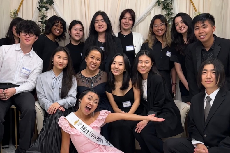 A large group of young adults, dressed in semi-formal black and white attire with name tags, pose together for a cheerful group photo at an indoor event. One woman in the front playfully extends her arms while wearing a sash that reads “Miss Universe.” Everyone is smiling and gathered closely, creating a warm, lively atmosphere.
