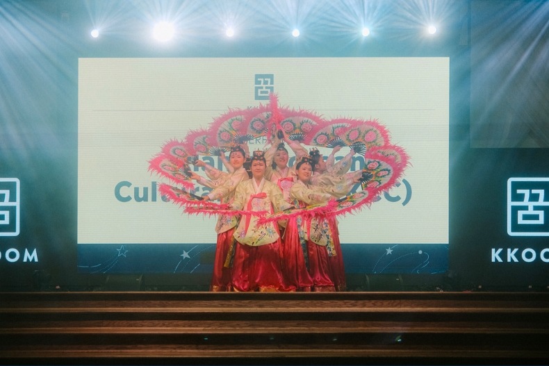 A group of performers in traditional Korean hanbok stand on stage holding pink and green fans arranged in a circular formation. Bright stage lights shine from above, and a large screen behind them displays part of the event program and the KKOOM logo. The dancers’ fans create a visually striking floral pattern.