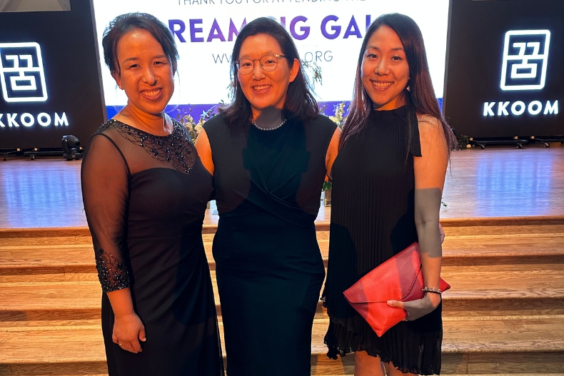 Three women dressed in elegant black evening attire stand together and smile at the camera at KKOOM’s Dream Big Gala. They are posed in front of a lit stage backdrop displaying the KKOOM logo and event signage. The atmosphere is formal and celebratory.