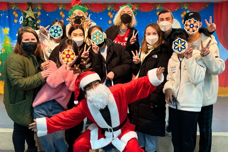 A group of volunteers and youth pose together at a festive holiday event, standing in front of a colorful Christmas-themed backdrop with trees, ornaments, and stars. Everyone is bundled in winter jackets, making peace signs and smiling behind face masks. Their faces are covered with decorative flower stickers. At the front, a person dressed as Santa Claus kneels playfully with arms outstretched, adding a fun and joyful energy to the group photo.