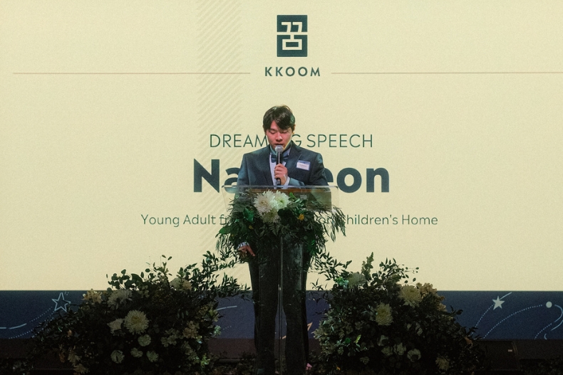 A young man stands at a podium delivering a speech on stage at the KKOOM Dream Big Gala. He holds a microphone and reads from his notes, surrounded by floral arrangements at the base of the podium. Behind him, a large screen displays the KKOOM logo and the words “Dream Big Speech” along with his name. The setting is formal and warmly lit, highlighting the significance of the moment.