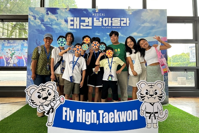 A group of students and two adults pose in front of a bright promotional backdrop for the musical “Fly High, Taekwon.” Everyone is smiling and making cheerful poses, with sticker icons covering each face. Two cartoon tiger mascots stand on both sides of the group, and artificial grass decorates the floor beneath them.