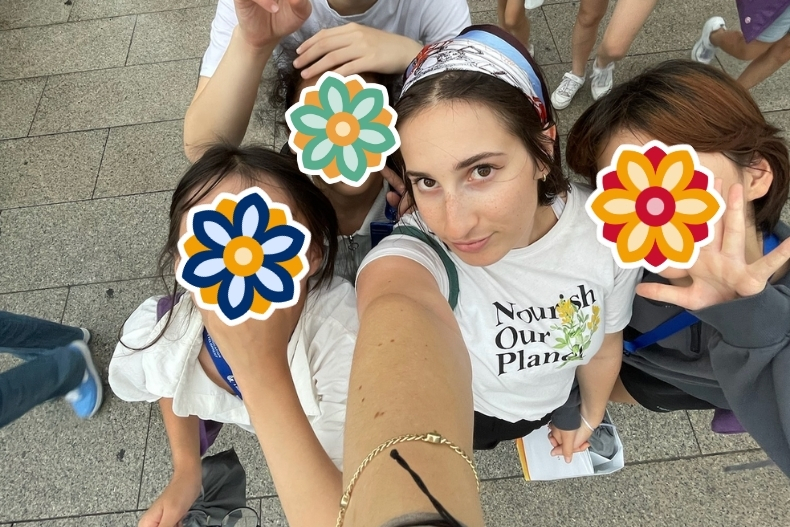 A group of girls pose playfully for a selfie with a volunteer. Their faces are covered with stickers as they smile and gesture toward the camera.