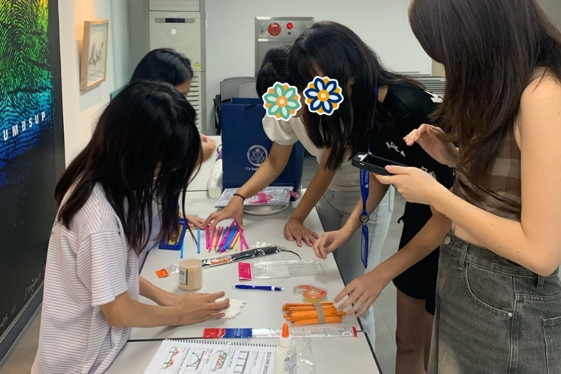 Girls participate in a hands-on STEM activity at a table, assembling small projects with volunteers assisting. Faces are covered with stickers.