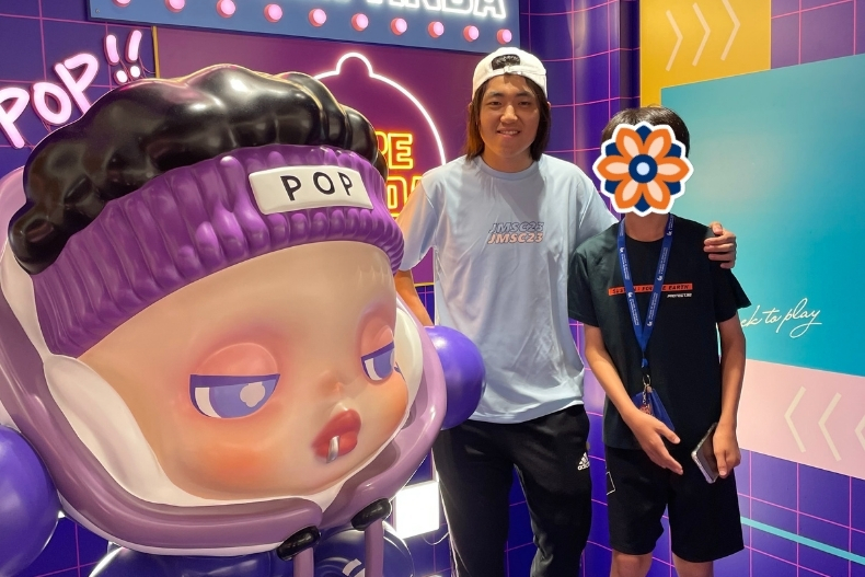 A volunteer stands next to a child whose face is covered with a flower sticker, posing in front of a colorful pop-art display featuring a large cartoon-style sculpture with purple hair and headphones.