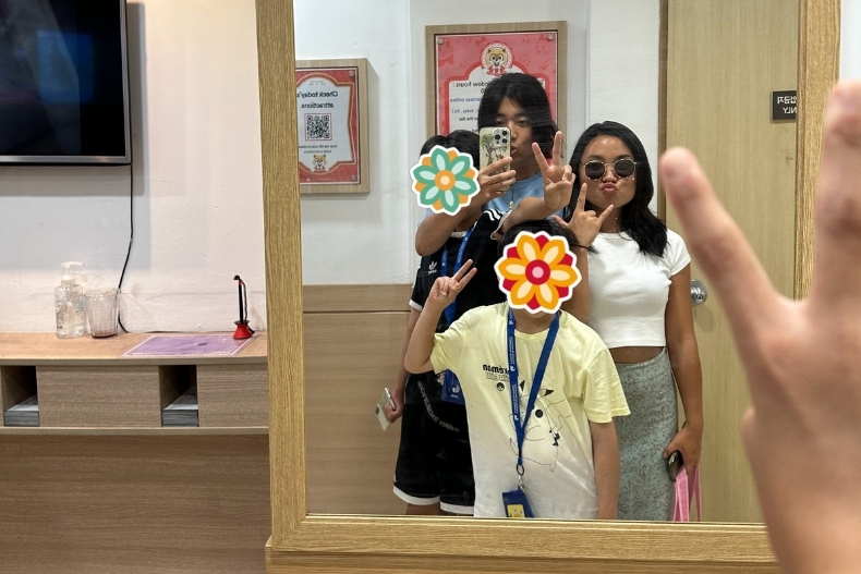 Two volunteers and a child with their face covered by a flower sticker pose together for a mirror selfie. One volunteer holds up a peace sign while the child stands in front, and the reflection shows a hand holding the phone.