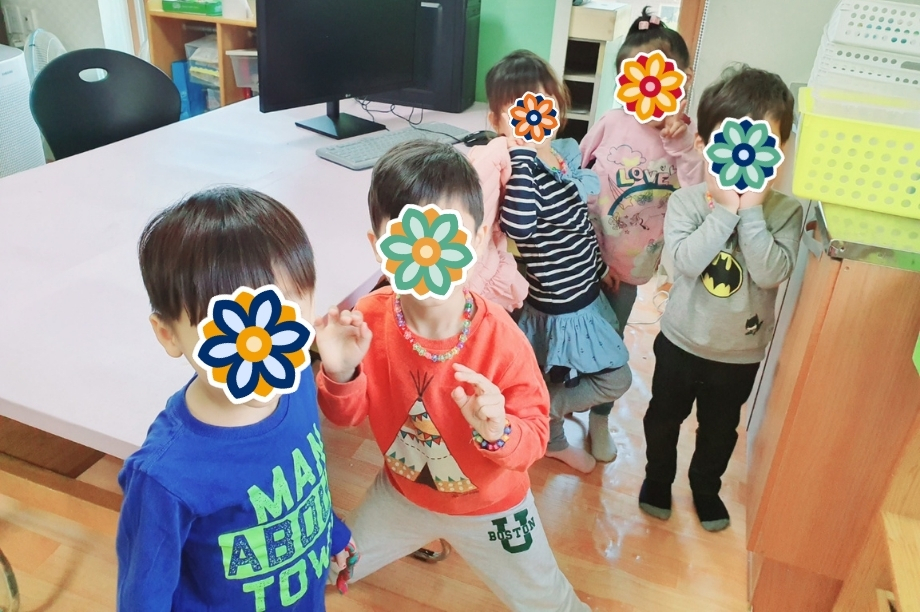 A group of young children stand in a classroom together, smiling and posing with peace signs and stickers covering their faces.