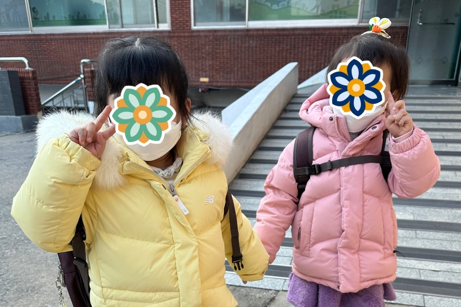 Two children wearing warm jackets and backpacks pose with peace signs outside a school building. Their faces are covered.