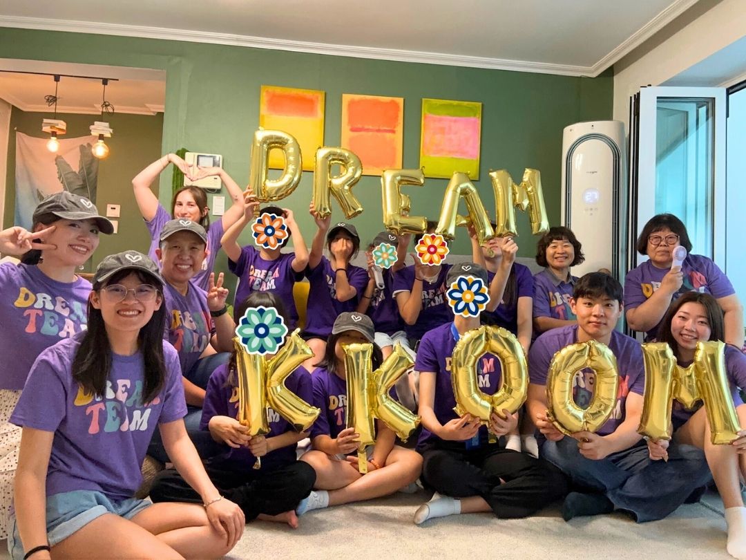 Children wearing purple shirts at a KKOOM camp, holding balloons spelling "DREAM KKOOM."