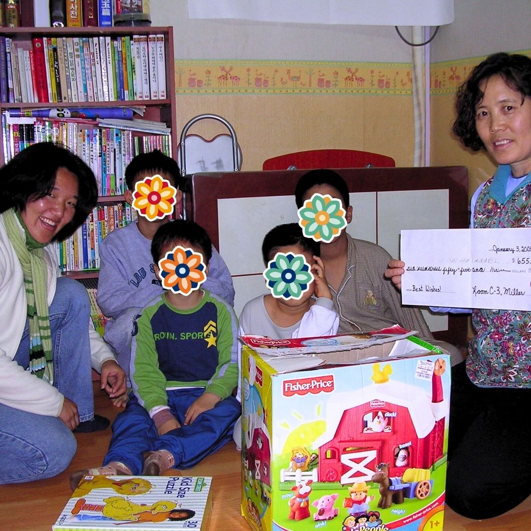 House mom holding a large donated check while sitting with the children and KKOOM’s co-founder during a holiday event.