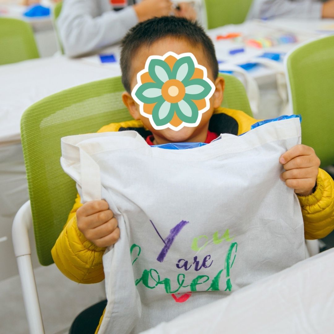 Child holding a handmade fabric bag with the message ‘You are loved’ during a KKOOM activity.