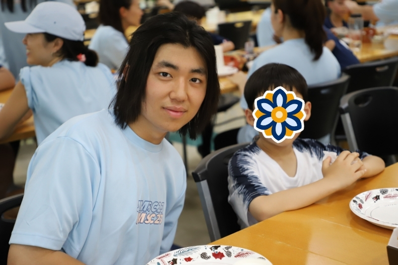 A volunteer wearing a light blue shirt sits at a cafeteria table and smiles at the camera. Next to him is a young child with their face covered by a flower sticker, sitting with hands folded on the table. Other participants can be seen seated in the background.