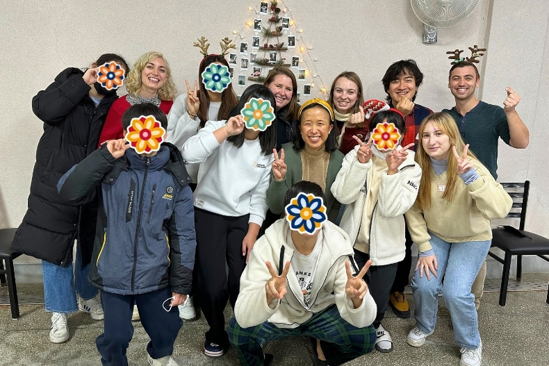 A group of volunteers and children pose together indoors, smiling and making peace signs. Several children’s faces are covered with flower stickers, and a decorated Christmas tree is in the background.